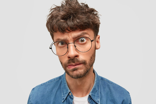 Close up shot of serious male boss with trendy hairdo looks in bewilderment at camera, frowns eyebrows, purses lips, has dark stubble, being annoyed with something. Negative facial expressions
