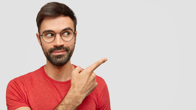 Horizontal shot of handsome bearded male with trendy hairdo, dressed in casual red t shirt, looks curiously asdie, points with index finger aside, shows free space for your advertising content