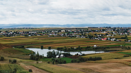Fototapeta na wymiar A typical natural landscape in the country of Suceava: fields, a small lake and a village of houses with red roofs, mountains on the horizon, Romania. Panoramic view.