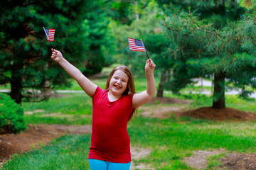 Fototapeta na wymiar Happy elementary age little girl smiles while holding American flag in her front yard on the Fourth of July. Independence Day, Flag Day