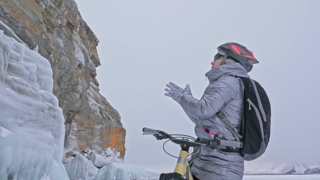 Woman is stay in bicycle on the ice. The girl is dressed in a silvery down jacket, cycling backpack and helmet. Woman is heating her hands with friction. Ice of the frozen Lake Baikal. The traveler is