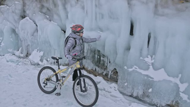 Woman is walking beside bicycle near the ice grotto. The rock with ice caves and icicles is very beautiful. The girl is dressed in silvery down jacket, cycling backpack and helmet. The tires on