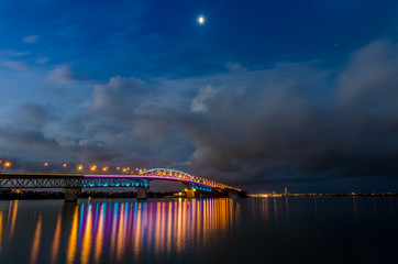 Auckland harbour bridge at night lit up in bright colours, with the moon.