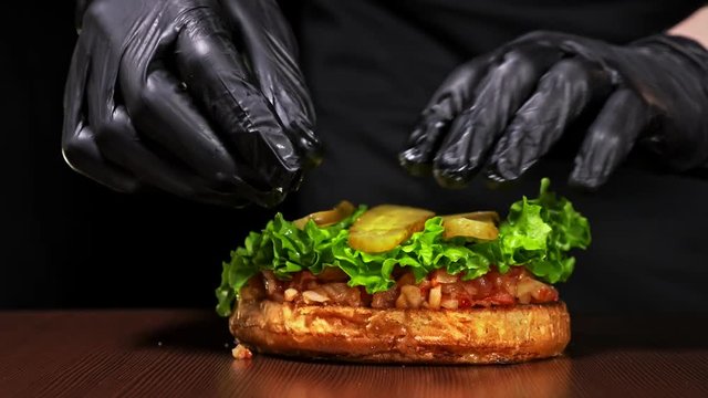 Burger is cooking on black background in black food gloves. Very luscious air bun and marbled beef. Restaurant where each burgers is cooked by hand. Put red onion and pickle cucumber on bun. Not made