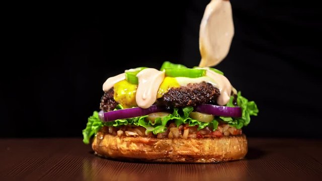 Burger is cooking on black background in black food gloves. Very luscious air bun and marbled beef. Restaurant where each burgers is cooked by hand. Sauce is poured onto burger. Not made ideal. Not