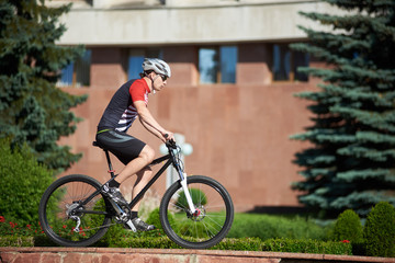 Man professional rider riding bike on street edging near flowerbeds and trees in front of red building. Sportsman training, getting ready for contest, improving skills. Concept of healthy lifestyle