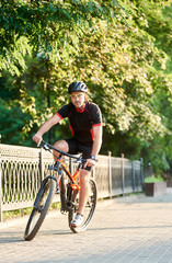 Fototapeta premium Athletic guy biker in cycling clothes riding on bike near green park. Concept of healthy lifestyle, sport advertising, outdoor activities