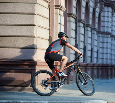 Back view of athletic cyclist in cycling clothes and protective gear riding bicycle in city center rushing and passing buildings. Sportsman training, exercising outdoors. Concept of healthy lifestyle