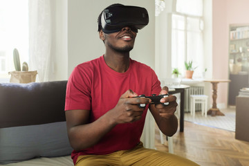 Young african american man playing game using VR glasses and wireless joystick, enjoying 360 degree virtual reality headset for gaming in contemporary cozy interior © Damir Khabirov