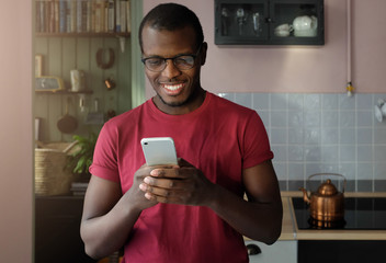 Closeup photo of African American man standing in cozy kitchen, looking at screen of cellphone,...