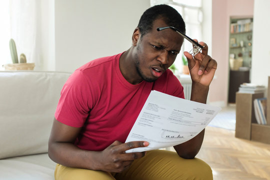 Shocked sad african young man surprised and stressed as read utility bill, holding eyeglasses