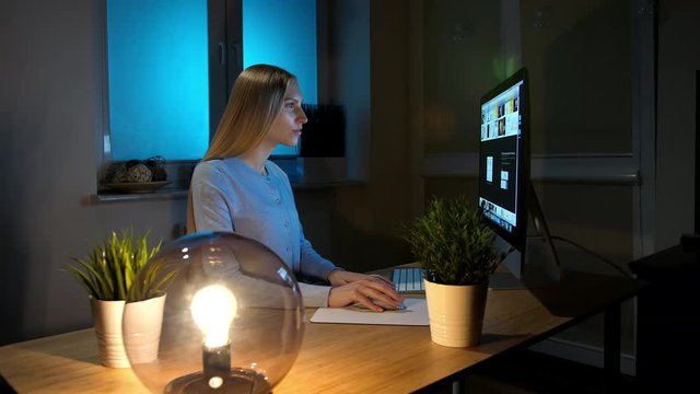 Woman looking attentively at computer at night. Relaxed beautiful blond female in warm home clothing sitting at wooden table lit by small sphere lamp and concentrating on browsing on computer.