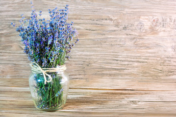 A photo of a bouquet of lavender in a glass vase with a neck bandaged with a rough rope on the left on a wooden background