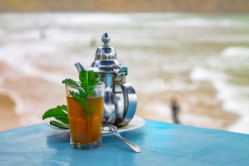 A glass of mint tea with an iron kettle, a sprig of mint and a large slice of sugar. A traditional...
