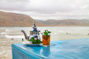 A glass of mint tea with an iron kettle, a sprig of mint and a large slice of sugar. A traditional Berber drink mint tea in the background of the Atlantic Ocean and mountains. Africa, Morocco, Agadir