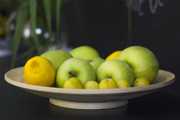 Fruits - lemons, green apples and yellow plums on a platter on a black wooden table