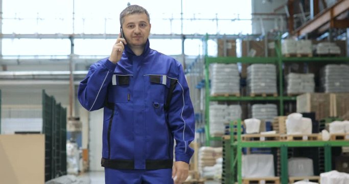 Engineer in in blue uniform is Walking Through Factory and talking on the mobile phone. front View