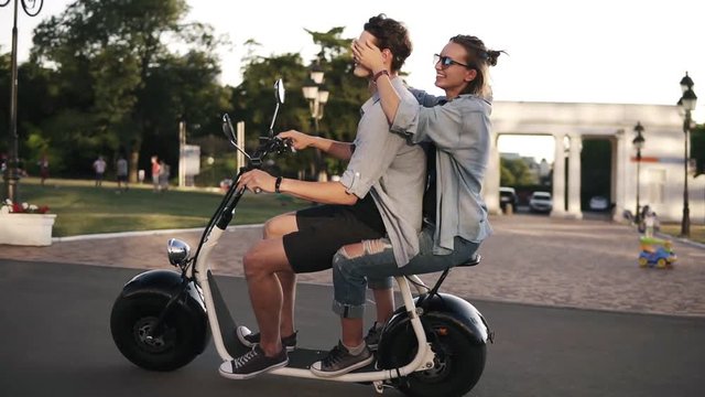 Young couple on dating riding an electronic minibike on the street. Wearing casual. Blonde lady closes his boyfriend's eyes while riding. Daytime