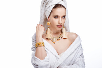 Fototapeta premium Young woman in a bathrobe and towel on her head, fashion golden jewelry, spa and care portrait, clean natural face, portrait on a background isolated