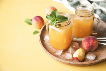 Cold summer peach drink with ice and mint in mason jars on a tray with fresh peaches on yellow background.