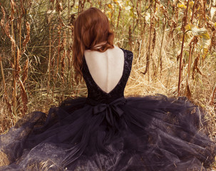red-haired beautiful girl sits with her back in black, long dress in the grass