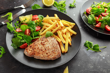 Fried Tuna Steaks on Black Plate with Fresh Green, Tomato Salad, lemon and french fries. healthy sea food