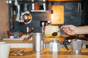 Mans hand with a spoon of ground coffee is filled with a geyser retro coffee maker on a wooden table.