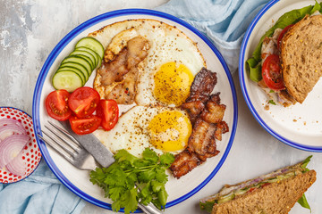 Fried eggs with bacon and a sandwich with meat, cheese and vegetables on white background, top view.