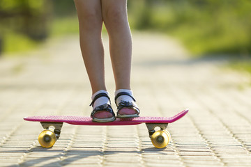 Girl slim legs in white socks and black sandals standing on pavement on plastic pink skateboard on...