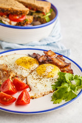 Fried eggs with bacon and a sandwich with meat, cheese and vegetables on white background. Delicious hearty breakfast.
