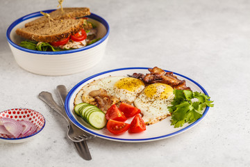 Fried eggs with bacon and a sandwich with meat, cheese and vegetables on white background. Delicious hearty breakfast.