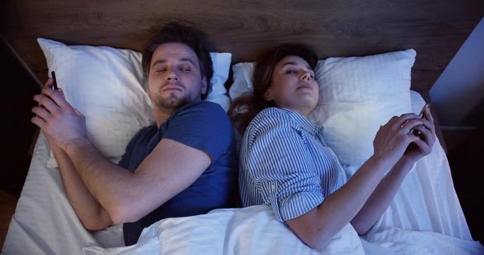 Close up of the married offended Caucasian man and woman lying in the bed back to back at night and taping on smartphones, then turning to each other for a second. Top view. Indoor.