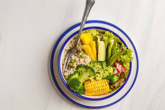 Vegan buddha bowl with brown rice, broccoli and vegetables in a white plate on a white background, copy space, top view.