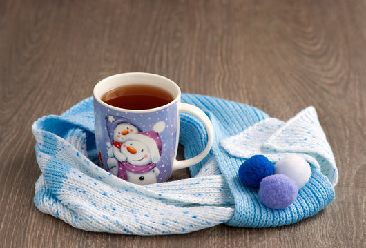 A mug of tea with a picture of a snowman and a scarf. A warm scarf wraps the hot tea on a cold winter day. New Year and snowman.