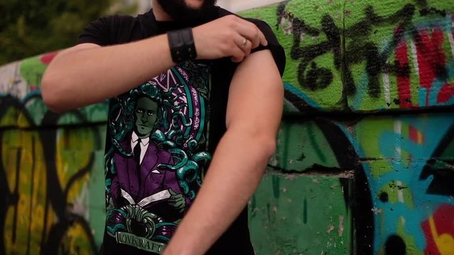 Young bearded strong man rolling up a sleeve on t shirt with bright print with wall with graffiti on the background