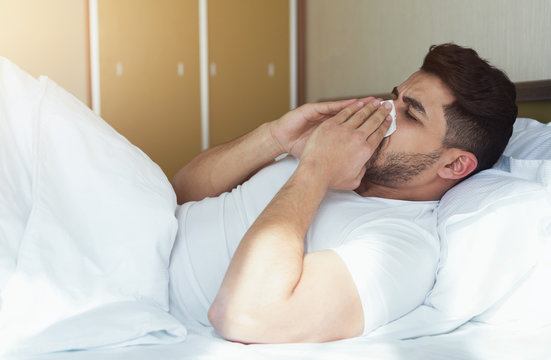 Young man sneezing and covering nose with tissue lying on bed