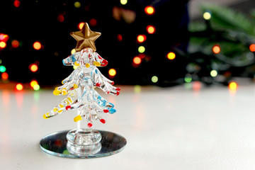 A small souvenir Christmas tree made of glass on the background of twinkling Christmas lights, bokeh effect. Empty space for text. 