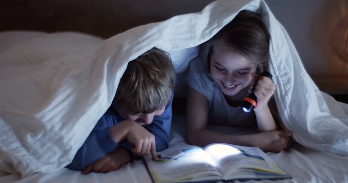 Close up of the cute little kids with a flashlight reading a book with excited faces under the blanket at night. Inside.