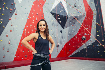 Portrait of happy fit girl with harness on her waist after successful ascent, rise, in climbing club studio over painted artificial rock background.