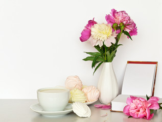 Romantic still life with fresh peonies in a vase, open book, a cup of milk drink and vanilla marshmallow on a light table. Free space for text
