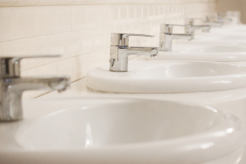 Many Closed chrome-plated faucets stand in a row in a public toilet. The room is clean and bright.