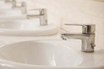 Many Closed chrome-plated faucets stand in a row in a public toilet. The room is clean and bright.