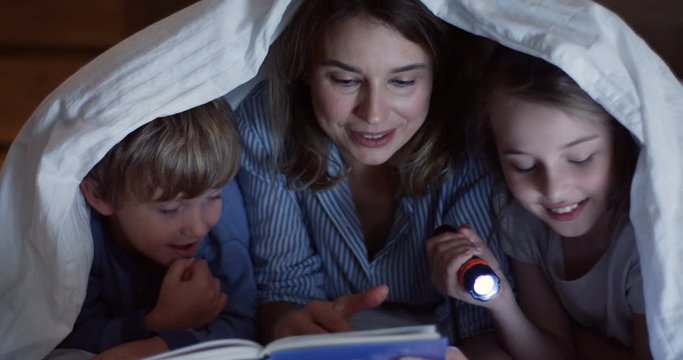 Close up of the caucasian beautiful mother and her cute small children with a flashlight reading the book and flipping pages under the blanket at night. Indoor.