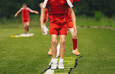 Obraz premium Young Athletes Training with Football Equipment. Football Speed Training with Ladder. Young Footballer Jumping in Red Sportswear at Training Session on Grass Soccer Field