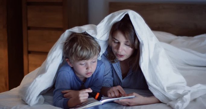 Portrait of the pretty caucasian woman reading a book for his small cute son while lighting with a little lantern under the blanket at night. Close up. Indoor.
