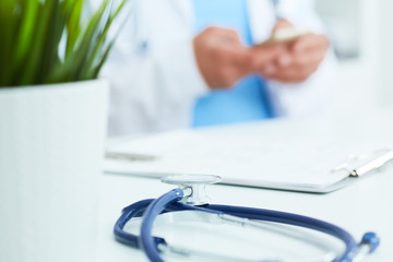 Blue stethoscope lies on the table in the foreground and male doctor hands texting on a smart phone in background.