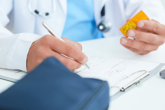 Male physician medicine doctor or pharmacist sitting at worktable, holding jar or bottle of pills in hand and writing prescription on special form.