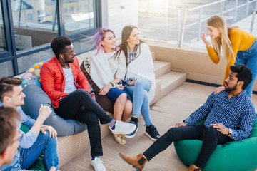 Young creative multiethnic people gesturing and discussing something with coworkers while sitting at the office lounge zone on soft poufs