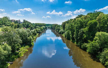 Fototapeta na wymiar Watercourse of Luznice river in Tabor city, Southern Bohemia, Europe. Scenic landscape with water surface and mirroring a blue sky with white clouds and banks with lush tree vegetation.