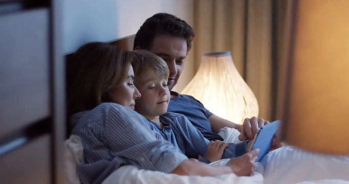 Cute little boy sitting among his parents in the bed in the evening. They watching something on the tablet computer with excited faces and talking about it. Indoors.